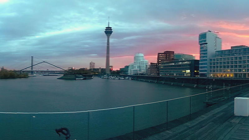 Example image show a cityscape at dusk with modern buildings, a river, and a prominent tower. The sky is painted with hues of pink and blue, reflecting on the water. The scene is calm and picturesque, capturing the essence of a serene urban evening.