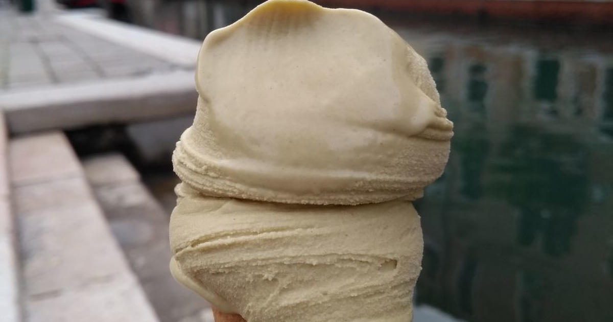 Hand holding an ice cream cone with a scoop of creamy ice cream by a canal lined with colorful buildings in a Venice.