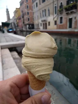 Hand holding an ice cream cone with a scoop of creamy ice cream by a canal lined with colorful buildings in a Venice.